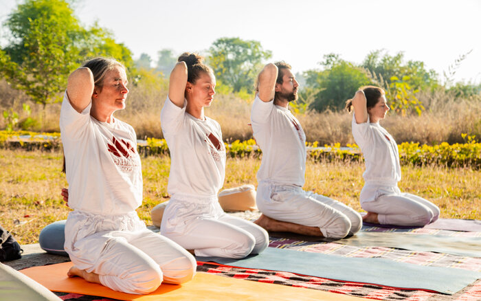 YTT students seated in Vajrasana practicing a shoulder opening pose Yoga students practicing a seated shoulder opening stretch in an outdoor setup
